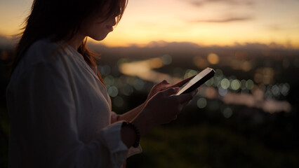 A woman uses her smartphone for social media communication during sunset with city lights in the...