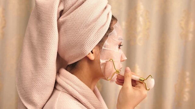 Woman in bathrobe with towel on head applying facial mask and massaging face with jade roller &mdash; skincare and beauty routine