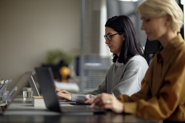 Caucasian young adult woman working on laptop in modern office, sitting beside blurred middle aged Caucasian woman using computer, both focused on tasks, professional environment