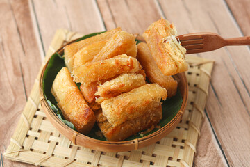 Person using fork to hold a golden fried cassava (Singkong Goreng), served warm and crisp in a traditional bamboo basket lined with banana leaf on a wooden mat. A beloved Indonesian comfort snack. 