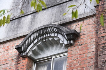 Authentic architectural detail of ornate arched window pediment with textured light material, fan grooves and dark curved vertical decorations above metal sliding window on red brick wall.