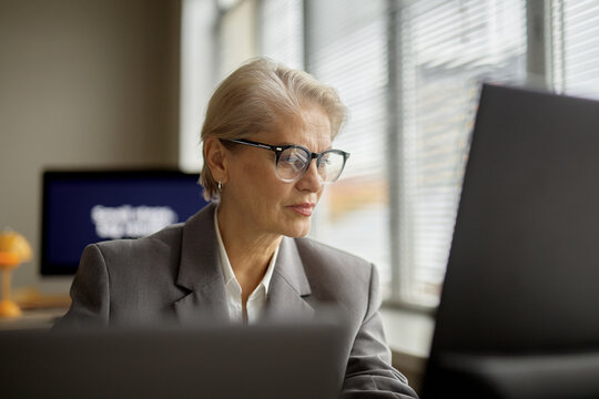 Senior Caucasian woman working at computer in modern office, wearing glasses and business suit, focusing on screen, sitting at desk with window blinds in background - Powered by Adobe
