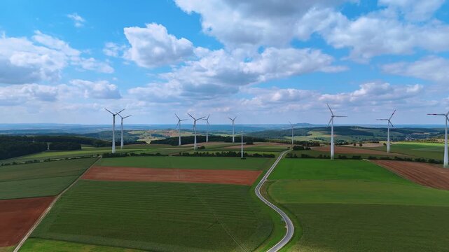 Wind farm over cultivated fields with cloudy sky. Drone landscape view of multiple wind turbines on green fields and brown soil in summer countryside.