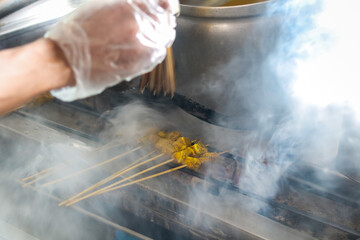 A person grills Sate or Satay Padang skewers over hot charcoal at a stall. The sizzling meat infuses with smoky flavor, showcasing the traditional process of this Indonesian street food dish.