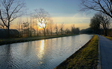 A tranquil sunrise over a calm canal, with trees lining the water's edge, creating a peaceful atmosphere.