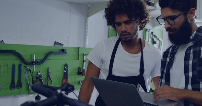Two bike mechanics wearing aprons using laptop in workshop with handlebars, pegboard and shelving