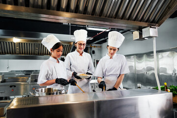 Three professional chefs in a commercial kitchen collaborate over a dish, wearing uniforms and hats...