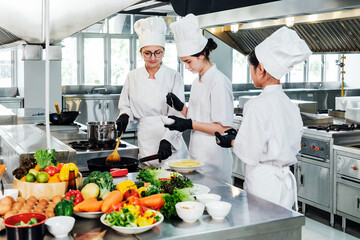Three chefs in white uniforms and hats cook in a professional stainless steel commercial kitchen, preparing dishes at the stove with fresh vegetables and ingredients arranged on the counter.
