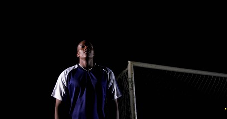 Standing soccer goalkeeper wearing jersey on field at night, gazing skyward with goal post and net
