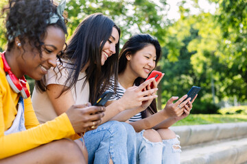 Three young multiracial girls using mobile phone sitting outside in summer. Social media and...