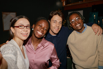 Group of Black and Caucasian young adults smiling and posing together for selfie, all looking directly into camera indoors