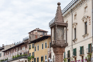 Stone column with carved Madonna relief stands in Verona’s main square, framed by frescoed Renaissance facades and pastel historic buildings