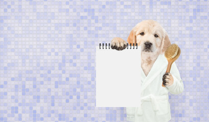 Cute Golden retriever puppy wearing bathrobe holding shower brush and showing blank notepad in it paws. Empty space for text