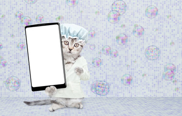 Cute kitten wearing shower cap and bathrobe standing on hind legs at bathroom and showing big smartphone with white blank screen in it paw. Empty space for text