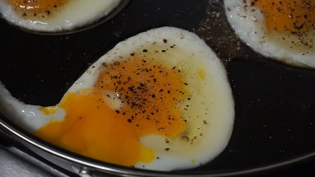 Close-up of fried sunny-side-up eggs in a pan, yolks sprinkled with pepper, visible whites and oil forming patterns during cooking