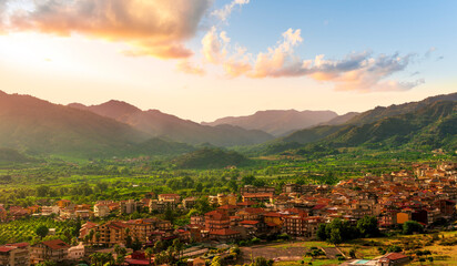 Obraz premium wonderful landscape of sunset valley town with beautiful fields, gardens, green plantations among great mountains and scenic evening cloudy sky on background of landscape