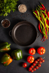 Top view of fresh vegetables and spices around an empty cast iron grill pan on dark stone table. Perfect food background for culinary design, restaurant menu, recipe blog, or healthy cooking concept.