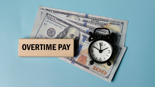 Concept of Overtime Pay with US dollar bills, a wooden block reading OVERTIME PAY, and an alarm clock, symbolizing extra work compensation and time equals money.