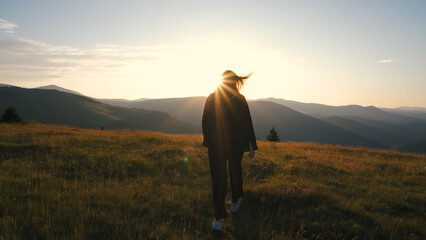 Woman walking in grassy landscape during sunset with mountains in background, capturing serene outdoor moment