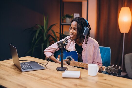 Young African American man with dreadlocks wearing headphones playing guitar and recording a podcast in his home studio