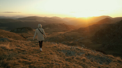 Woman walks through a scenic mountain landscape at sunset, enjoying the peacefulness of nature and capturing the beauty of the horizon