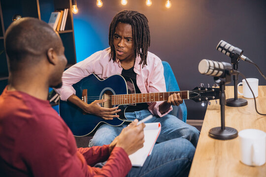 Two African American men creating content for their music podcast, one playing guitar and singing, the other taking notes