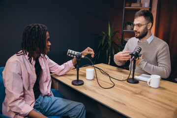 Two male podcasters sitting at a wooden table, talking and gesturing during a podcast recording session