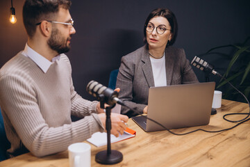 Professional podcasters recording audio episode, sharing insights while speaking into microphones and working with laptop in soundproof studio environment
