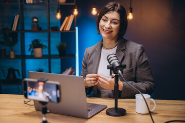 Happy female podcaster speaking into microphone and recording audio podcast at home studio using laptop and smartphone