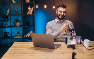 Happy podcaster speaking and clapping hands during a live podcast recording in his studio with laptop and smartphone