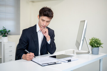 Fototapeta premium Asian businessman holding pen thinking as looking at clipboard on computer table in company's office