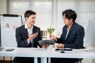 Asian businessman holding clipboard while talking to coworker at working table in company's office.