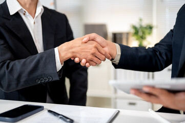 Close up of businessman holding tablet shaking hands with client at working table in company office.