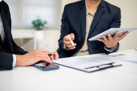 Close up of businessman holding pen and tablet while talking to coworker at table in company office. - Powered by Adobe