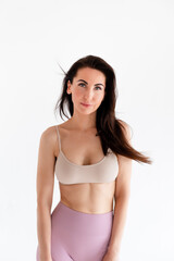 Fototapeta premium Sports woman standing in activewear with abs muscles, looking at camera isolated on white background. Caucasian 30s fitness girl wears beige top, pink leggings. Happy fit smiling brunette, blue eyes.