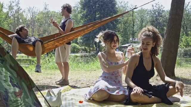 A group of diverse friends enjoys a summer picnic in a lush park, applying sunscreen and relaxing in a hammock. They share laughter and good times, making memories together outdoors.