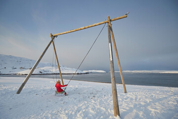 A girl in a red jumpsuit swings on a large swing against the backdrop of the non-freezing northern sea and snow in winter