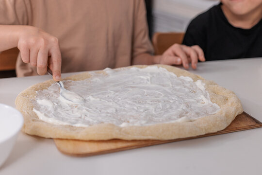 Children preparing pizza at home and spreading white sauce on dough, concept of family cooking, learning and creativity.