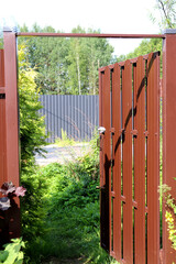 The metal gate of a rural house is open to the street, road, trees, and the fence of a neighboring house on a sunny summer day - color vertical photo