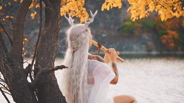 silhouette fantasy woman fairy faun plays a flute, sitting under tree creating magical music. Myth sexy beauty girl fae back rear view, blonde hair long white dress deer antlers on head. river rock