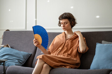 Woman at home suffering from overheating, sitting on sofa in living room holding hand paper fan for...
