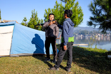 Two Happy Asian Men Celebrating With a Cheers With Their Mugs at Their Campsite by a Tranquil Lake