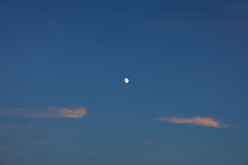 Serene moon hangs high in clear dusk sky, flanked by soft wispy clouds showing hint of pink hue from sunset's scattering sunlight.