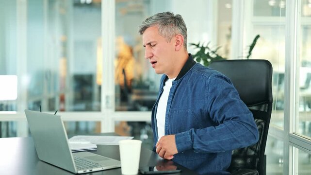 Tired businessman, dressed in a denim jacket and white t-shirt, experiences painful back discomfort while working on a laptop at his office desk. The sedentary office work causes strain and pain