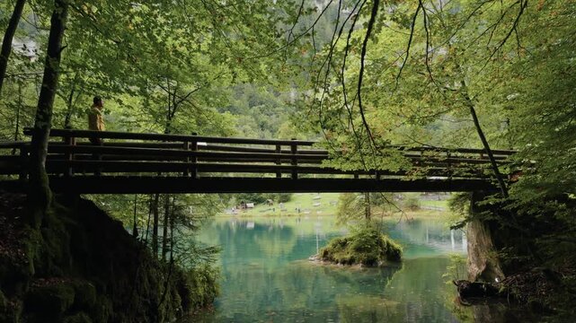 Traveler in a yellow jacket walking on a wooden bridge at Blausee in Switzerland, with the turquoise alpine lake in the background. Scenic summer adventure lifestyle in nature