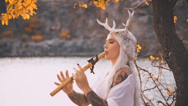 Portrait fantasy woman fairy faun plays a flute, sitting under yellow tree creating magical music. Myth happy sexy beauty face girl fae, long blonde hair white dress deer antlers on head. river rock 
