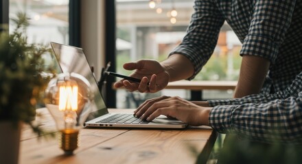Person using laptop on wooden desk with light bulb and plant near window.