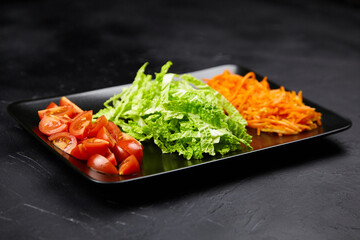 Freshly sliced tomatoes, vibrant green lettuce, and shredded carrots arranged on a black platter, showcasing colorful vegetables for healthy meal preparation and culinary presentation