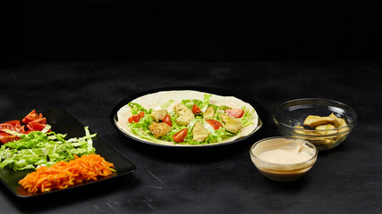 Freshly prepared salad with lettuce, tomatoes, and carrots served on a black background, accompanied by a bowl of sauce and crispy snacks, showcasing vibrant colors and textures