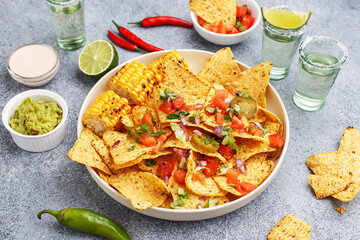 Mexican nachos corn chips served with chopped tomatoes, onion and greens, marinated jalapeno, grilled corn, various dips and tequila shots on gray concrete background. Party snacks. Side view